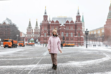 A beautiful young girl in a pink jacket walks along Manezhnaya Square in Moscow during a snowfall and blizzard. Snowblowers are working in the background.