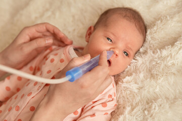 Little baby lying on bed on fluffy blanket, faceless mother using nasal aspiration for taking care of her new born child. Kid with her mom, kid wearing sleeper with polka dots.