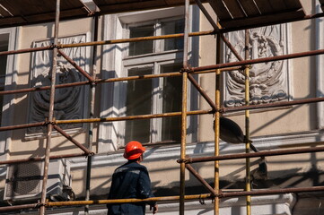 Man builder in orange construction helmet working from scaffolding to renovate historic building wall with ornate sculptural relievo details. Historic building restoration in progress