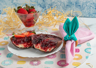 Two slices of Easter egg with a juicy filling of strawberry jam, biscuit, chocolate and cream, next to a pink napkin with napkin ring similar to rabbit ears and a bowl of strawberries