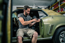Young Man Removing Interior Trim Panels From An Old Vintage Car From The 60s Or 70s In His Home Garage. Tools Are Seen Around.