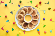 Plate Of Small Round Pancakes With Colorful Party Topper Flags On Split Three Color Layered Paper Background. Top View, Bold Vibrant Colors, Orange, Green, Yellow. Jar Of Honey. Creative Food Concept.