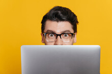 Amazed Excited Caucasian Young Adult Guy With Glasses Peeking Out From Behind Laptop, Looking Surprised At Camera While Standing Against Isolated Orange Background, Close-up