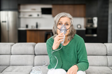 sick senior gray-haired woman sitting on the couch in a living room, wearing an oxygen mask to treat