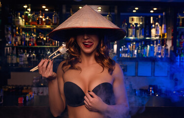 Sexy young female model standing in black bra with asian hat on the head and sushi sticks in her hands with a piece of roll 