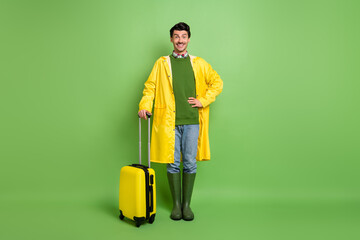 Poster - Full size photo of young happy cheerful positive man in yellow coat hold baggage isolated on green color background