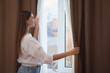 © New Africa - Woman opening elegant window curtains in room