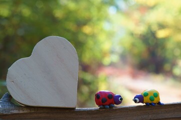 Wall Mural - Two ladybugs made of plasticine in the forest on a colored background. Next to it is a wooden heart.