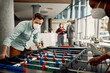 © Drazen - Businessman with face mask playing table football with colleague while taking break from work in the office.