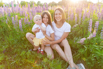 Wall Mural - Young mother embracing her kids outdoor. Woman baby child and teenage girl sitting on summer field with blooming wild flowers green background. Happy family mom and daughters playing on meadow.