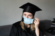 © Iryna - Graduate girl in medical mask at coronavirus covid-19 period. Woman with master degree in black graduation gown and cap. Distance learning online. Study at home. Graduation from college.