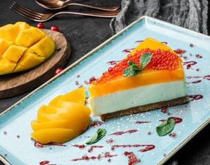 Creamy mango cheesecake decorated with red berries and mint on a large plate and dark background, close up view. Summer desserts and sweets