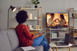 © Studio Romantic - Woman enjoying good romcom serial in the evening. Single African American lady sitting alone at home, drinking coffee and watching romantic love story on soap opera entertainment TV channel, back view