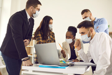 Diverse business team in face masks reduce COVID-19 infection risk. Multiethnic colleagues using laptop brainstorming idea discussing financial strategy sitting at desk. New normal office lifestyle