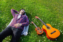 Young Man With Sunglasses Lying On The Grass Field With Flowers Around, Acoustic Guitar And Ukulele Around Him