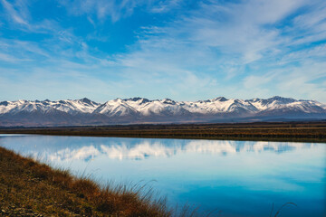 Wall Mural - Snow covered Southern Alps mountain range near Twizel reflected in the still water in the canal flowing out of Lake Rautaniwha under some wispy cloud