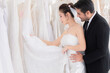 © Rakchanok - Wedding shopping lifestyle Concept, beautiful Asian girl show dress to groom making decision for wedding ceremony, man help woman choose bridal dress in fitting room, happy couple spent time together