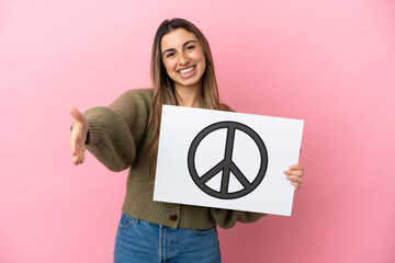 Wall Mural - Young caucasian woman isolated on pink background holding a placard with peace symbol making a deal