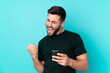© luismolinero - Caucasian handsome man isolated on blue background with phone in victory position
