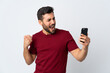 © luismolinero - Young handsome man with beard using mobile phone isolated on white background celebrating a victory