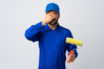 Poster - Painter man holding a paint roller isolated on white background covering eyes by hands. Do not want to see something