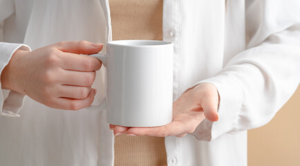 Wall Mural - Female hands are holding a mockup of a white empty mug or cup of coffee tea, for your design and advertising, business concept on a beige background, Scandinavian style. Woman in white shirt