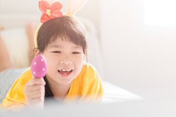 Happy easter day.Asian child girl in rabbit ears holding eggs.Online worship in easter holidays with laptop.Kid child online with bunny hat, easter egg hunt online at home.Lockdown, covid19, medical.