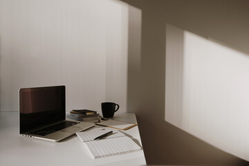Wall Mural - Minimalist home office desk workspace. Laptop on table with coffee cup, paper sheet, stationery against white wall. Work, business background. Sunlight shadow on the wall.