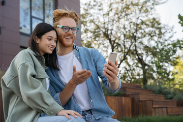 Wall Mural - Two smiling friends using mobile phone shopping online. Beautiful asian woman and handsome caucasian man communication together, watching videos on smartphone sitting in park 