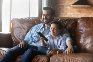 Canvas Print - Smiling young Caucasian father and little son relax on sofa in living room watch TV together. Happy dad and small boy child rest on couch on family weekend enjoy television. Entertainment concept.