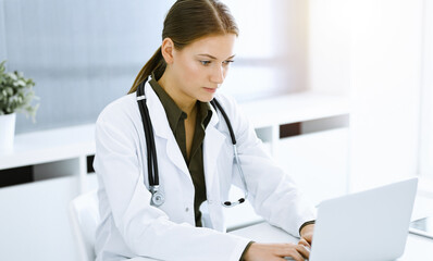 Wall Mural - Woman-doctor typing on laptop computer while sitting in sunny hospital office. Data in medicine and healthcare