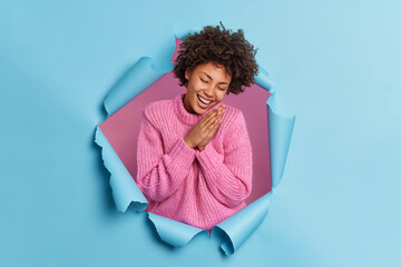 Wall Mural - Sincere optimistic curly haired woman keeps palms pressed together chuckles from joy expresses natural emotions wears knitted sweater poses through paper ripped background. Happiness concept