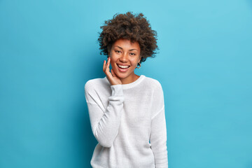 Wall Mural - Happy dark skinned charmming woman with curly hair smiles broadly touches face enjoys soft skin has natural beauty expresses positive emotions dressed incasual jumper isolated on blue studio wall