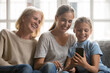 © fizkes - Smiling three generations of Caucasian women relax at home using modern smartphone gadget together. Happy little girl child with young mother and senior grandmother look at cellphone screen laughing.