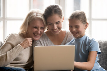 Wall Mural - Happy three generations of Caucasian women look at laptop screen laugh talking on video call online. Smiling girl child with young mother and older grandmother use computer relax together at home.