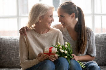 Wall Mural - Happy caring young female congratulate greet overjoyed older Caucasian mother with birthday anniversary at home. Smiling loving adult grownup daughter make surprise present gift and flowers to mom.