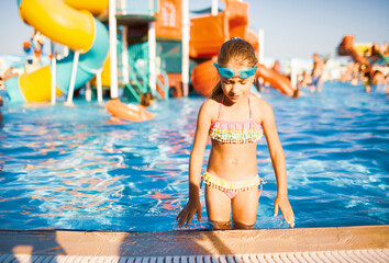 Adorable girl in goggles for swimming standing in the pool and sprinkles raising her hands up