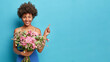 Happy African American woman with curly hair holds bouquet of flowers wears festive dress indicates at copy space shows advertisement or promotion enjoys spring and holidays isolated on blue wall
