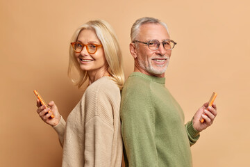 Wall Mural - Senior woman and man stand backs to each other use modern cellphones stand backs to each other wear spectacles and casual sweaters isolated over brown background. Family technology age concept