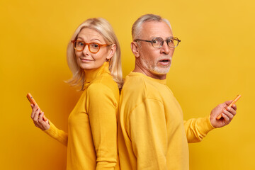 Wall Mural - Two elderly female and male friends stand back to each other wear optical glasses casual jumpers use modern gadgets for online communication type text messages isolated over yellow background.