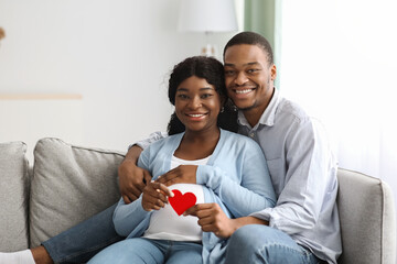 Wall Mural - Cheerful expecting couple sitting on couch, holding red heart