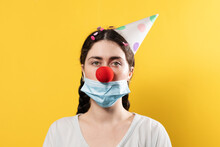 April's Fool Day Concept. Portrait Of A Woman In A Paper Cap And A False Red Nose With A Protective Mask. Yellow Background. Copy Space. New Normal