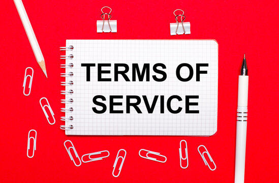 On a red background, a white pen, white paper clips, a white pencil and a notebook with the text TERMS OF SERVICE. View from above