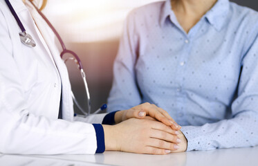 Unknown woman-doctor is holding her patient's hands to reassure a patient, while sitting together at the desk in the sunny cabinet in a clinic. Female physician is using a clipboard and a stethoscope