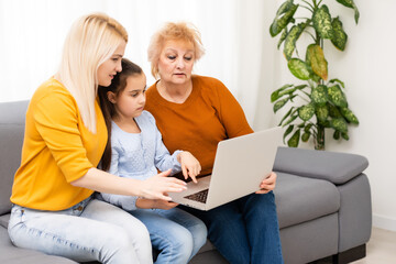 Wall Mural - Granny, her daughter and granddaughter are using a laptop and smiling while sitting on sofa at home