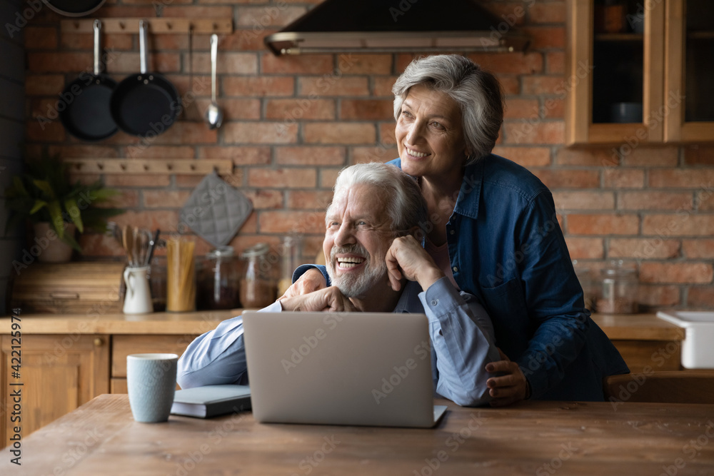 Looking forward together. Loving aged spouses pensioners spend time together at country cottage kitchen use laptop dreaming. Bonding senior couple husband wife hug look at distance imagine good future - obrazy, fototapety, plakaty 