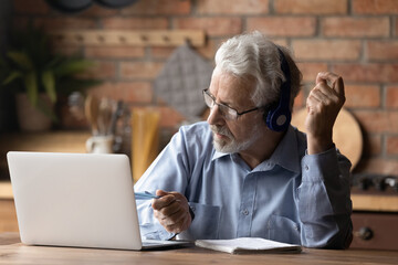 Third age student. Focused old man in headphones study from home listen to webinar training on laptop take notes. Older male worker take part in virtual briefing discuss work with colleagues online