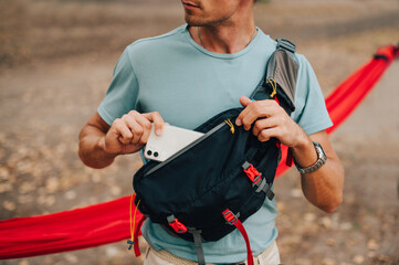 A young man in a T-shirt and a waist bag on his shoulder puts a smartphone in his pocket, close up