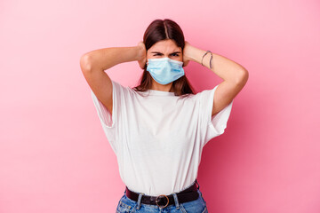 Young caucasian woman wearing a mask for virus isolated on pink background covering ears with hands trying not to hear too loud sound.