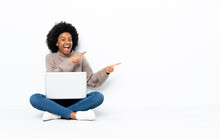 Young African American Woman With A Laptop Sitting On The Floor Surprised And Pointing Side